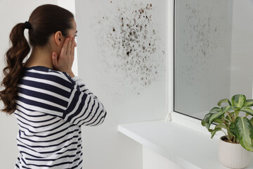 Shocked woman looking at affected with mold window slope in room