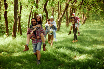Group of young people, friends walking in forest, going hiking on warm sunny day. Girls sitting on boys backs. Concept of active lifestyle, nature, sport and hobby, friendship, leisure and fun - obrazy, fototapety, plakaty