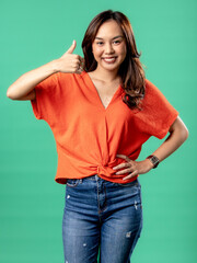 A portrait of an Asian woman wearing an orange shirt, expressing joy and giving a thumbs-up, isolated on a green background.