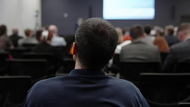 A man in a blue sweater and glasses listens to a lecturer at a business meeting