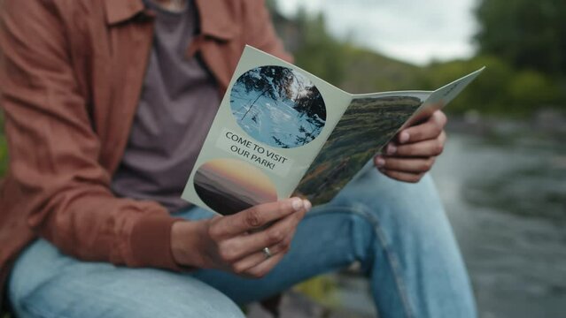 Shallow focus of unrecognizable person holding and reading tourist brochure with river background