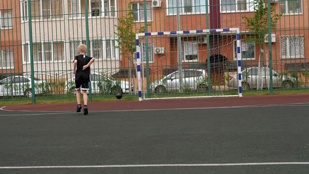 A boy trains with a soccer ball on a rubber-coated playground in the courtyard of a modern multi-storey building. The child hits the ball on goal.