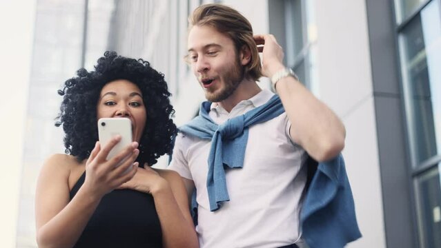 Cute young interracial couple looking at smartphone screen while get notification message and celebrate online win success reading great news feel amazed happy outdoors Triumph Lucky Wow concept