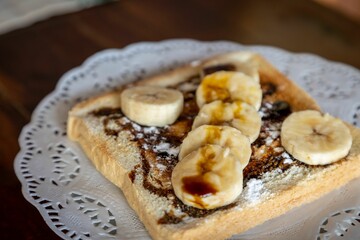 Horizontal close-up of a toast with chocolate and banana pieces on a white plate on a wooden table