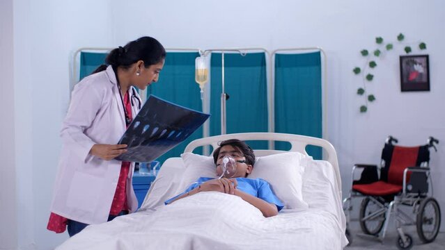 A young lady doctor consulting a sick young boy on the hospital bed - X-ray  MRI  medical report  CT-scan. An Indian boy wearing an oxygen mask is resting in the hospital room - a medical condition...