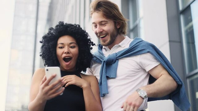 Cute young interracial couple looking at smartphone screen while get notification message and celebrate online win success reading great news feel amazed happy outdoors Triumph Lucky Wow concept