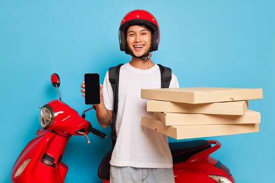 Young pizza guy in red helmet stands on blue background near red motor bike with smile, holds boxes with pizza in one hand and black phone in another, delivery concept, copy space