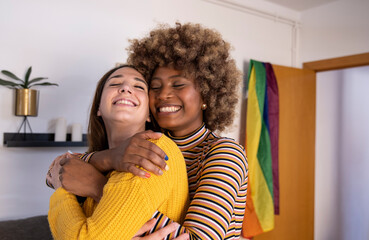 Beautiful and cheerful multiracial lesbian couple hugging at their apartment. Two lovely and happy homosexual women embracing each other with their eyes closed at home. - obrazy, fototapety, plakaty