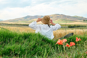 Rear view of woman in straw hat sitting in poppies meadow and admiring golden wheat field in hot...