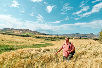 Naklejka na ściany i meble Middle-aged bearded man walking in golden wheat field in hot summer sun and blue sky with white clouds with mountains hill landscape in background. Leisure activity outdoors.