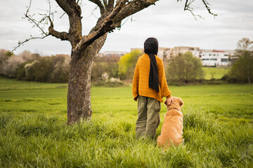 young muslim girl standing with a dog, two best friends under a tree in a park while watching the...
