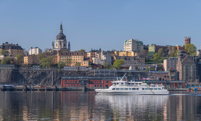 Obraz premium A modern commuting ferry leaving for the archipelago passing the cliffs of the district Södermalm part Sofo, a tranquil sunny summer day in Stockholm