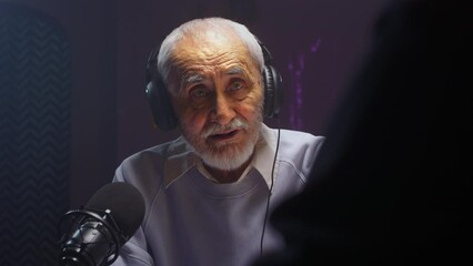 The old man creates a podcast in a professional studio. Senior male does online streaming interviews. Elderly man blogger speaking in microphone or make vlog. Recording audio content for social media