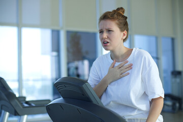 Tired exhausted young woman is training workout, running on treadmill in gym holding her head with hand, suffering from headache, feeling unwell, unhealthy during run 