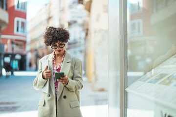 Beautiful mid adult woman walking and texting message on mobile phone outside business center....
