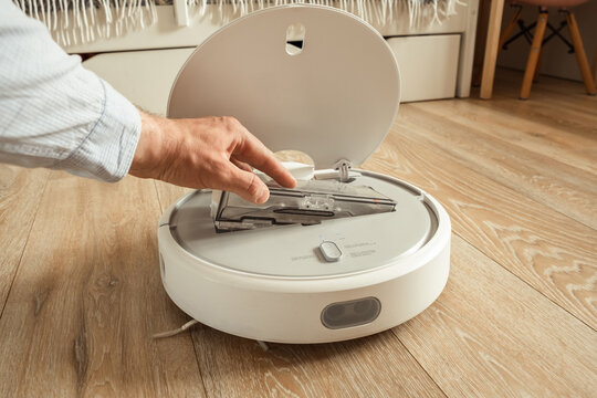 Robotic Vacuum Cleaner Working On Laminate Wood Floor In The Living Room. Man Removing Trash Bin Out Of The Automated White Vacuum Cleaner Robot With The Open Lid.