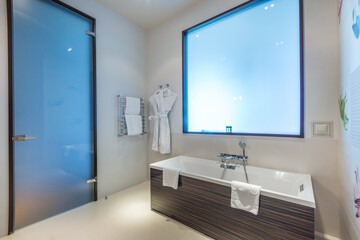Stylish design of the bathroom in the hotel, with a frosted glass door and a false window. Bath with shower, heated towel rail and bathrobes. - obrazy, fototapety, plakaty