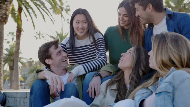 Happy group of millennial friends having fun sitting outdoors. Diverse college students smile and laugh together relaxing together at street. Friendship and youth lifestyle concept.