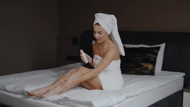 Pretty happy woman with towel on her head is sitting on the bed, puts creme on her leg, enjoying her time to relax. Beautiful caucasian woman relaxing on bed after shower in bedroom at home