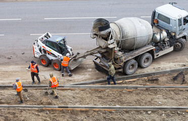 Team of road workers in orange signal vests transfer concrete from concrete mixer truck into mini loader bucket on road being repaired. Top view