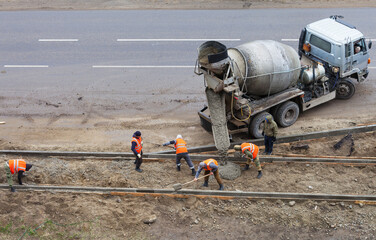 Team of road workers in signal orange vests receive concrete from concrete mixer truck and place...