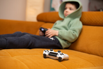 Cute 5 year old boy playing video game console while sitting on orange couch at home.	 - 605357214
