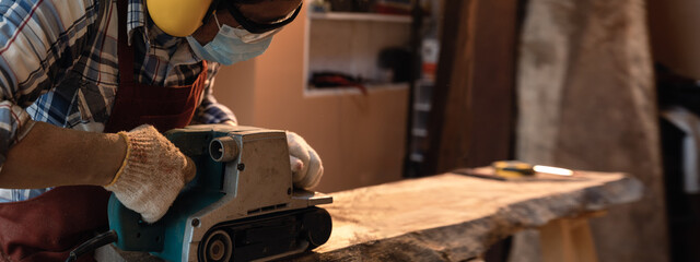 Carpenter use a hand-held electric sanding machine belt sander to level surface wood in carpentry workshop.