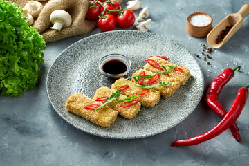 Breaded fried tofu with soy sauce and chili pepper in a plate. Gray background