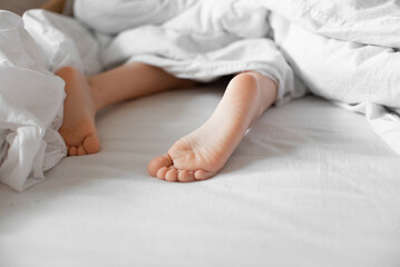 Obraz na płótnie Canvas Baby feet under white cotton bed linen. Morning natural light.