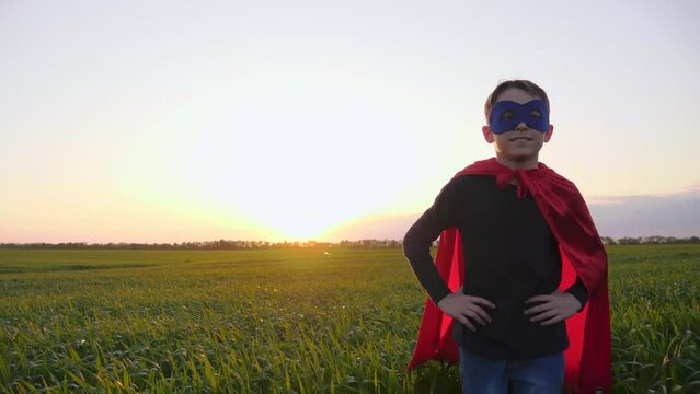 Portrait of a kid in a superhero mask and a red cape. A happy boy go a green meadow on the background of a beautiful sunset. Kids dreams