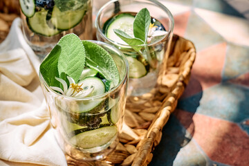 Refreshing summer drink - cucumber infused water with ice, sage, cucumber and lemon blossom on the table in the garden. Fresh healthy cold detox beverage. Fitness drink.