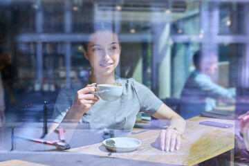 Young female inside cafe with cup of coffee, view through a glass window