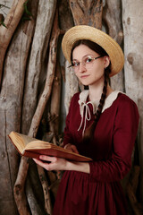 A young woman in a straw hat and round glasses is reading a book. Romantic style, boho, rustic atmosphere. A modest dress with a white collar