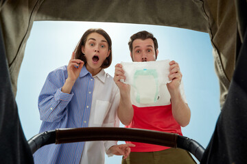 Young emotional parents, happy man and woman looking into baby carriage during walk on warm sunny...