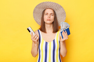 Sad unhappy woman tourist holding mobile phone tickets passport miss her journey wearing striped swimming suit posing isolated over yellow background displeased expression.