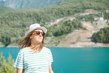 Fototapeta premium Smiling woman in hat and sunglasses with wild hair standing near mountains lake on background. Positive young woman traveling on blue lake outdoors travel adventure vacation.