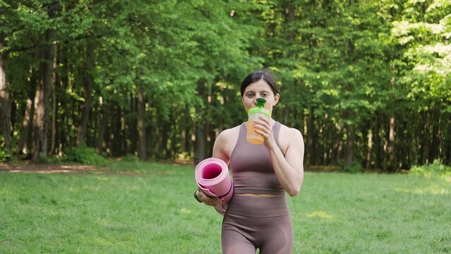 Sporty woman with yoga mat drinking sport nutrition with bcaa amino acids and vitamins from shaker, outdoors in summer park after morning workout, slow motion, healthy lifestyle concept