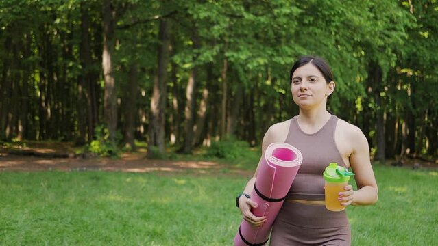 Sporty woman with yoga mat drinking sport nutrition with bcaa amino acids and vitamins from shaker, outdoors in summer park after morning workout, slow motion, healthy lifestyle concept