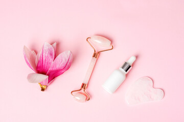 Obraz na płótnie Canvas Rose quartz crystal facial roller and gua sha scraper, face serum or organic cosmetic oil, magnolia flower on pink background. Facial massage kit for lifting therapy. Top view, flat lay