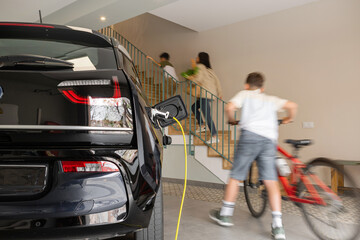 Plugged cable charging an electric family car in a particular garage while boy arrives with bicycle...