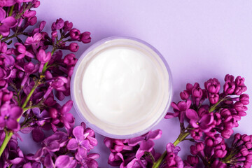 Obraz na płótnie Canvas Cream jar and lilac flowers on light purple background. Natural cosmetics, skin care concept. Top view, flat lay