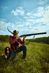 Portrait with male hunter wearing retro clothes with hat holding gun and shooting with his companion dog English springer spaniel over nature landscape background