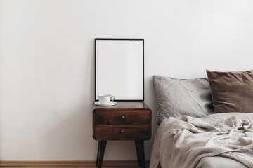 Vertical black picture frame mockup. Retro wooden mahogany bedside table, cup of coffee. Beige linen throw, brown velvet pillow in bed. White wall background. Scandinavian bedroom, elegant interior. - obrazy, fototapety, plakaty
