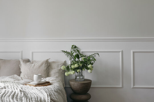 Breakfast in bed. Cup of coffee, wicker tray. Green bouquet of white viburnum, fern and solomons seal flowers. Modern boho night stand. Bedroom view. Beige pillows, linen blanket. Elegant moulding.