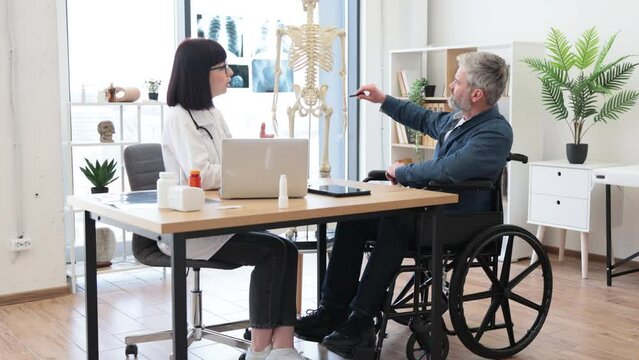 Full length view of female physician and man with disability examining skeleton model while staying in doctor's office. Attentive adults analyzing spinal cords for better awareness of pain source.