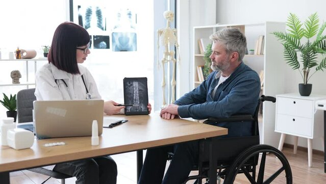 Attractive caucasian man taking pen for marking on tablet with x-ray image carried by radiologist in consulting room. Confident specialist discussing results of diagnostic tests with wheelchair user.