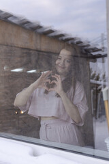 Fototapeta premium Photograph through glass. Young beautiful girl standing at the window and showing a heart