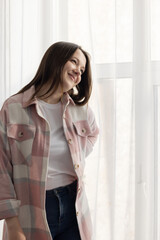 Young beautiful girl in plaid shirt standing at window with her hands propped up on her back