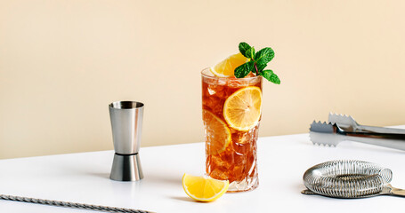 Long Island ice tea cocktail drink with vodka, rum, tequila, gin, liquor, lemon juice, cola and ice with lemon slice and mint in highball glass. Beige background, hard light, bar tools