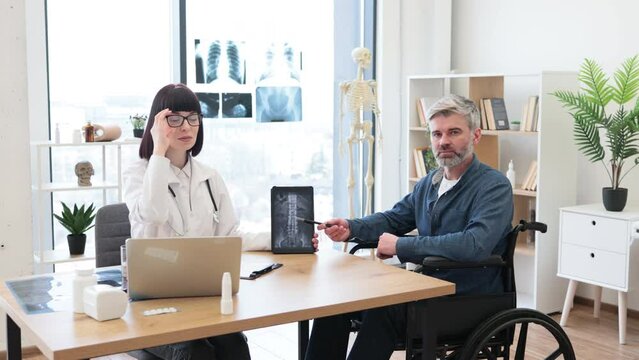 Full length portrait of female doctor and serious male patient with disability posing at office desk in clinic. Health professional giving recommendation about treatment due to CT scan images.